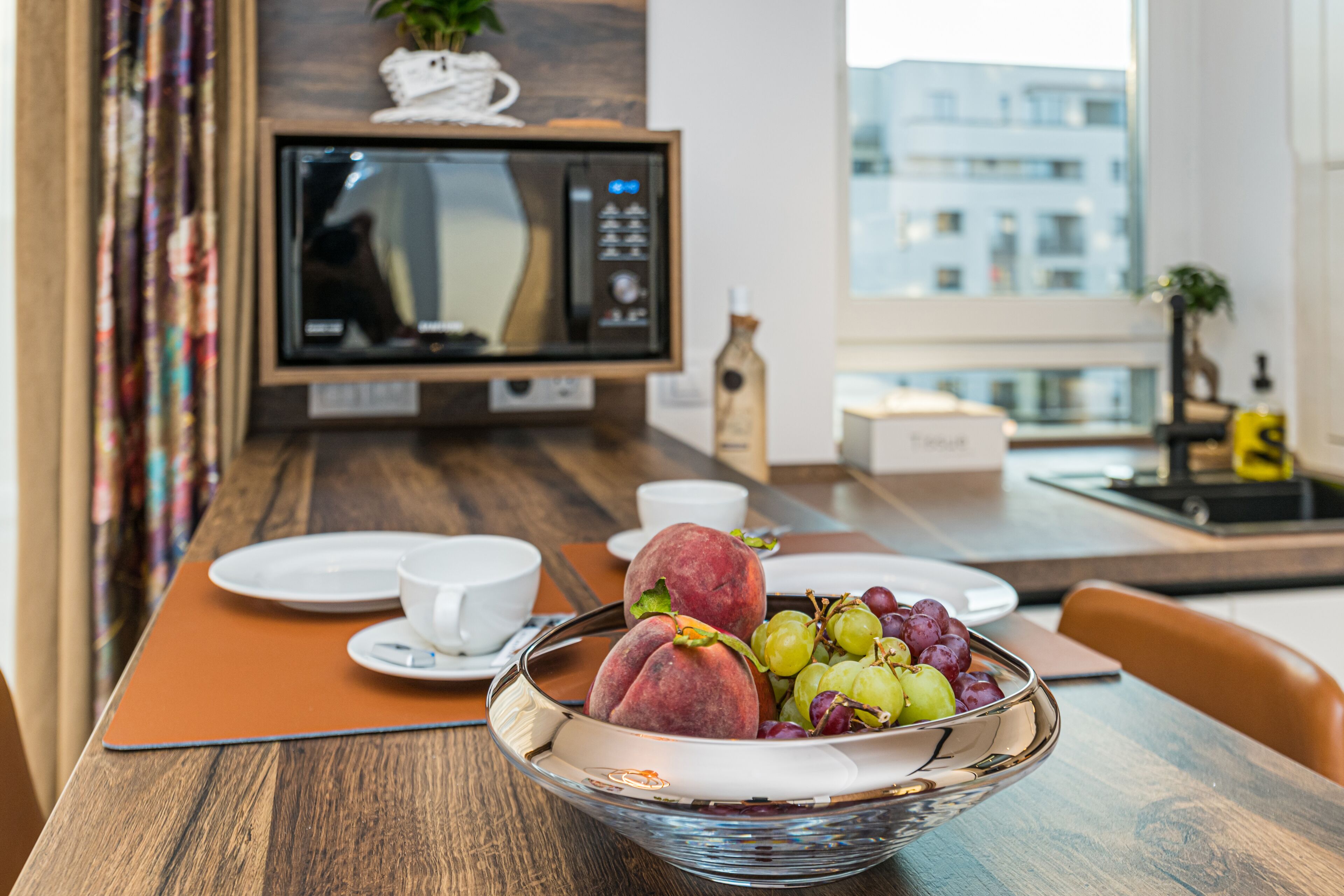 A modern kitchen with a microwave, a dining table with plates, cups, and a fruit bowl.