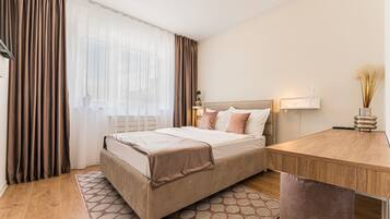 A hotel room with a bed, a desk, a chair, a window with curtains, and a bedside table with a vase.