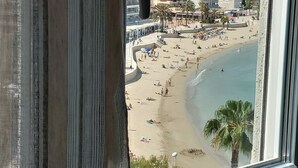 Vista a la playa desde una ventana con cortinas, mostrando una playa de arena, palmeras y un paisaje urbano al fondo.