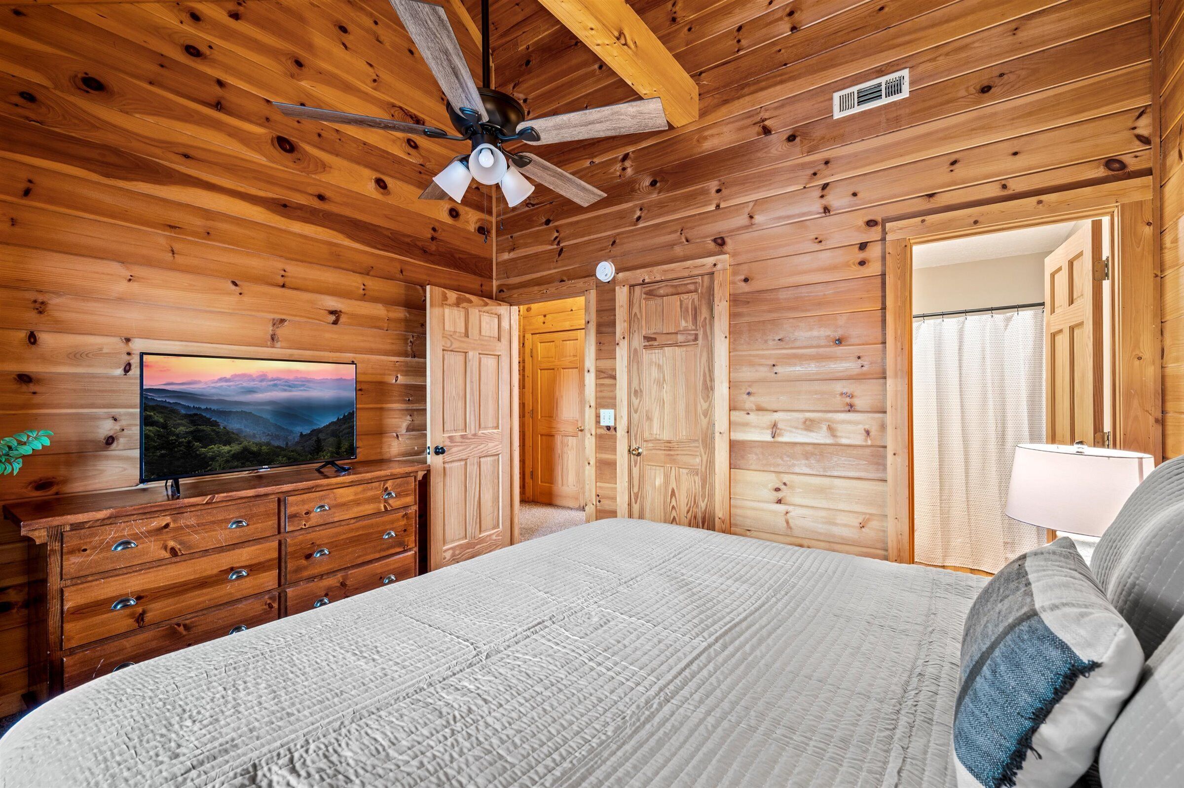 A wooden cabin bedroom with a bed, dresser, TV, and ceiling fan.