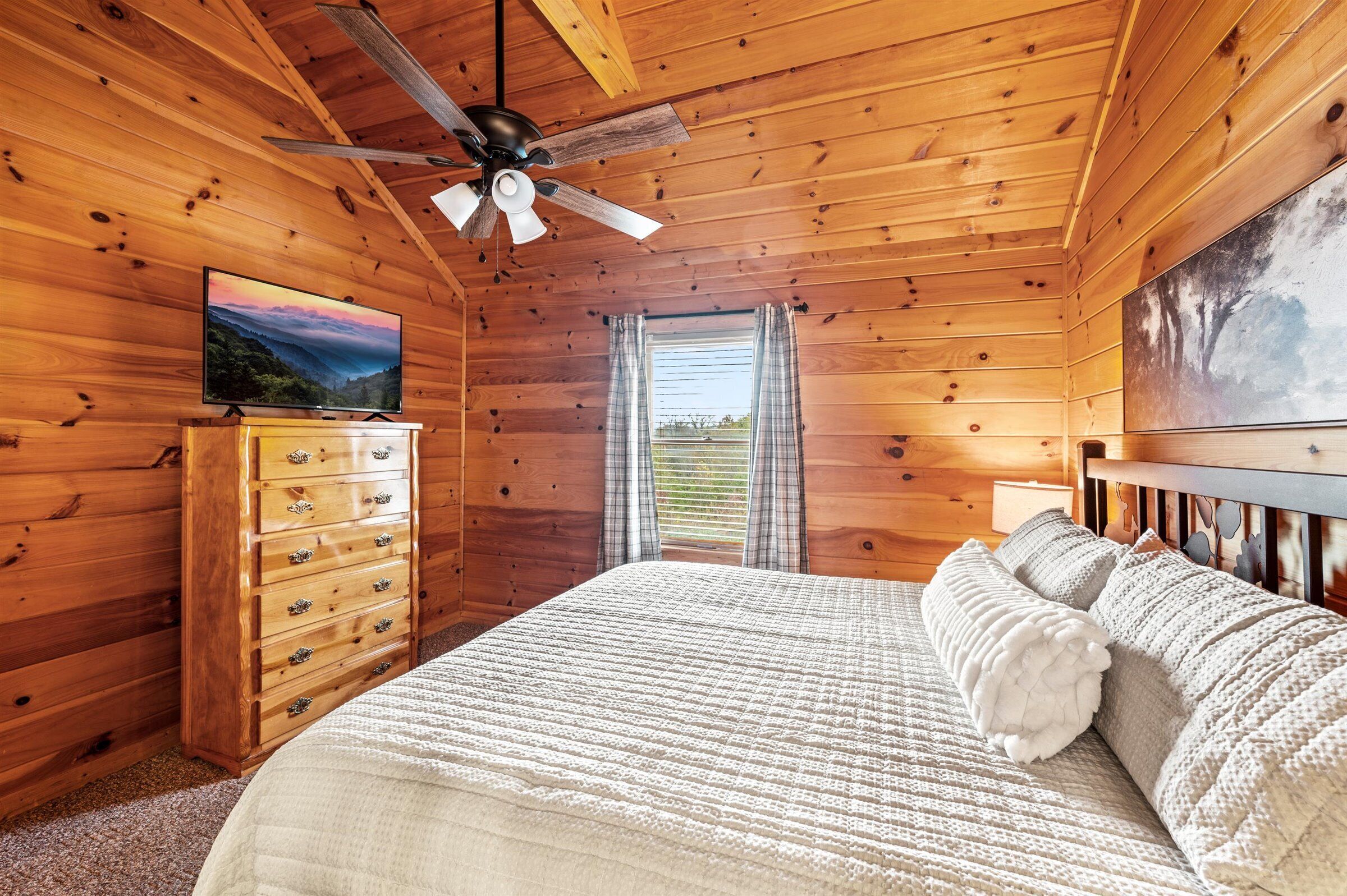 A wooden cabin bedroom with a bed, dresser, ceiling fan, and a painting on the wall.