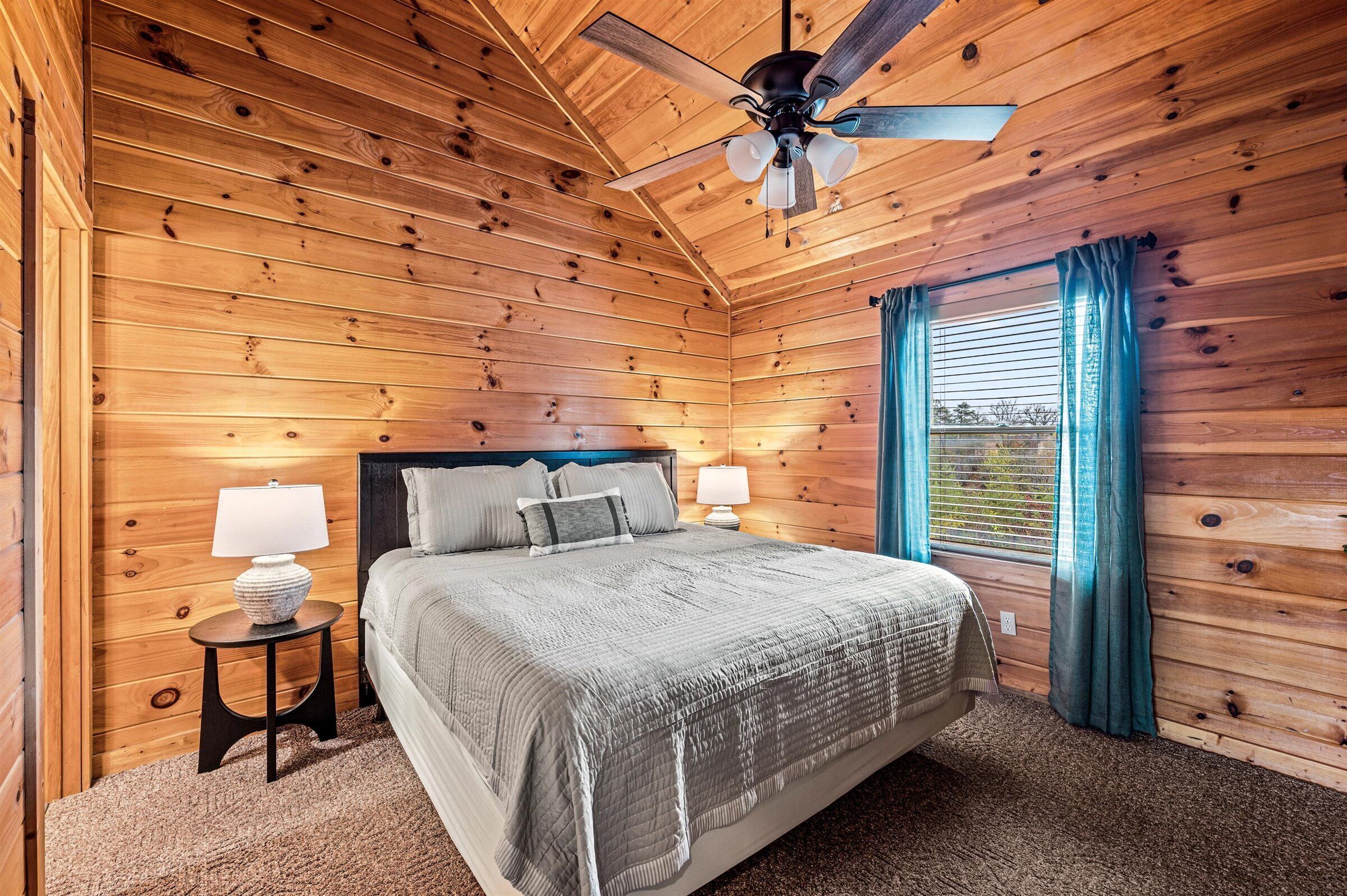 A wooden-paneled bedroom with a bed, two bedside lamps, a ceiling fan, and a window with curtains.
