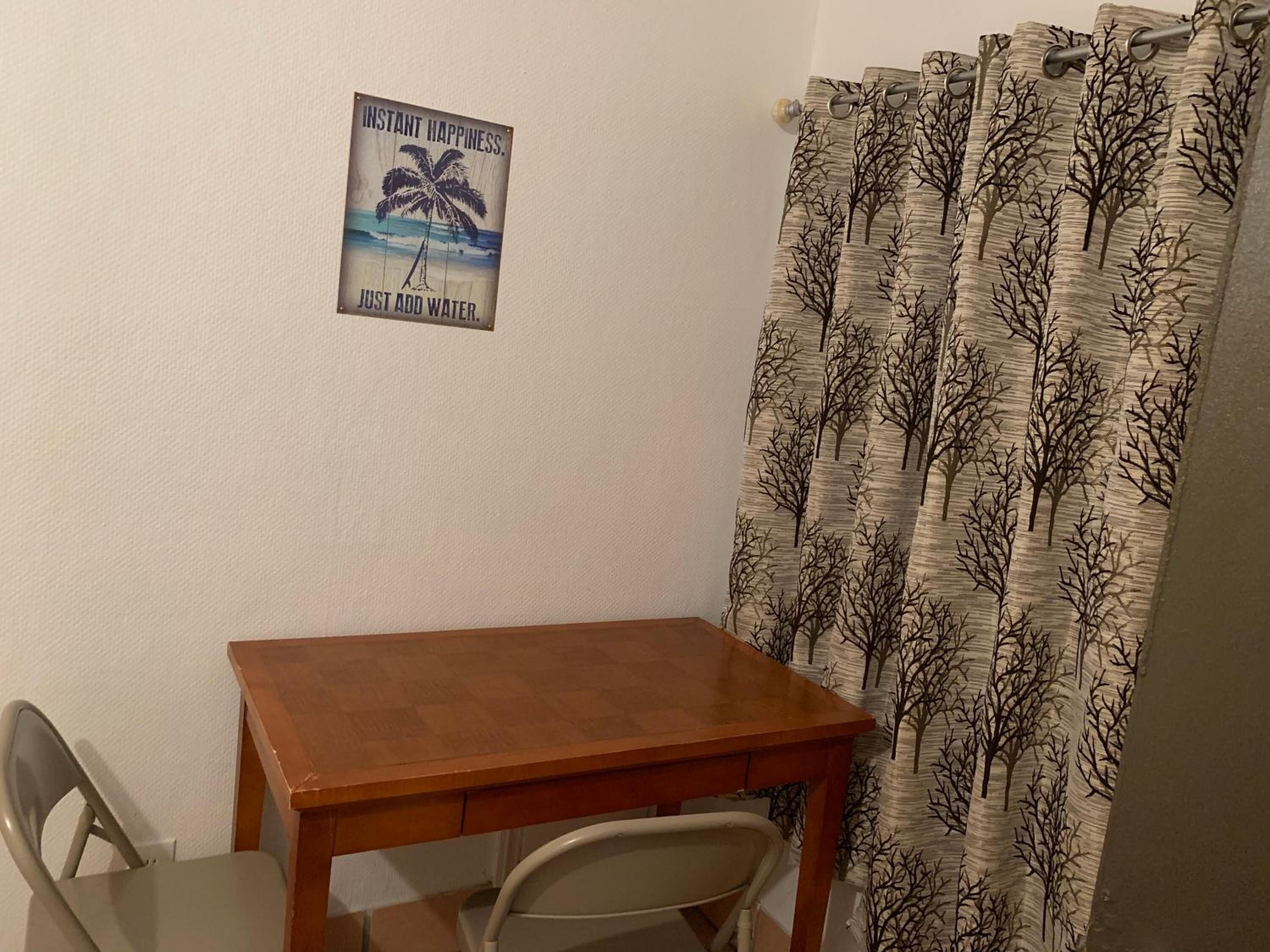 Une petite pièce avec une table en bois, une chaise, une affiche au mur et un rideau orné de motifs d’arbres.