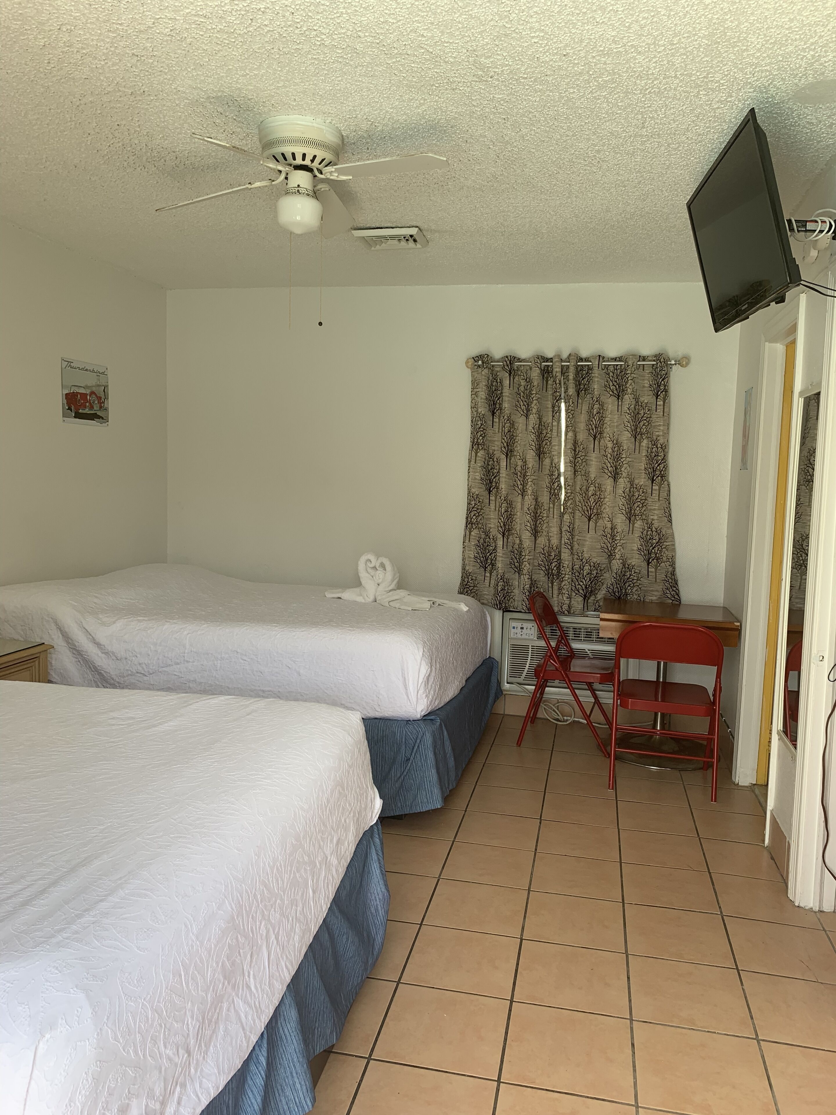 A room with two beds, a desk with a chair, a ceiling fan, and a TV mounted on the wall.
