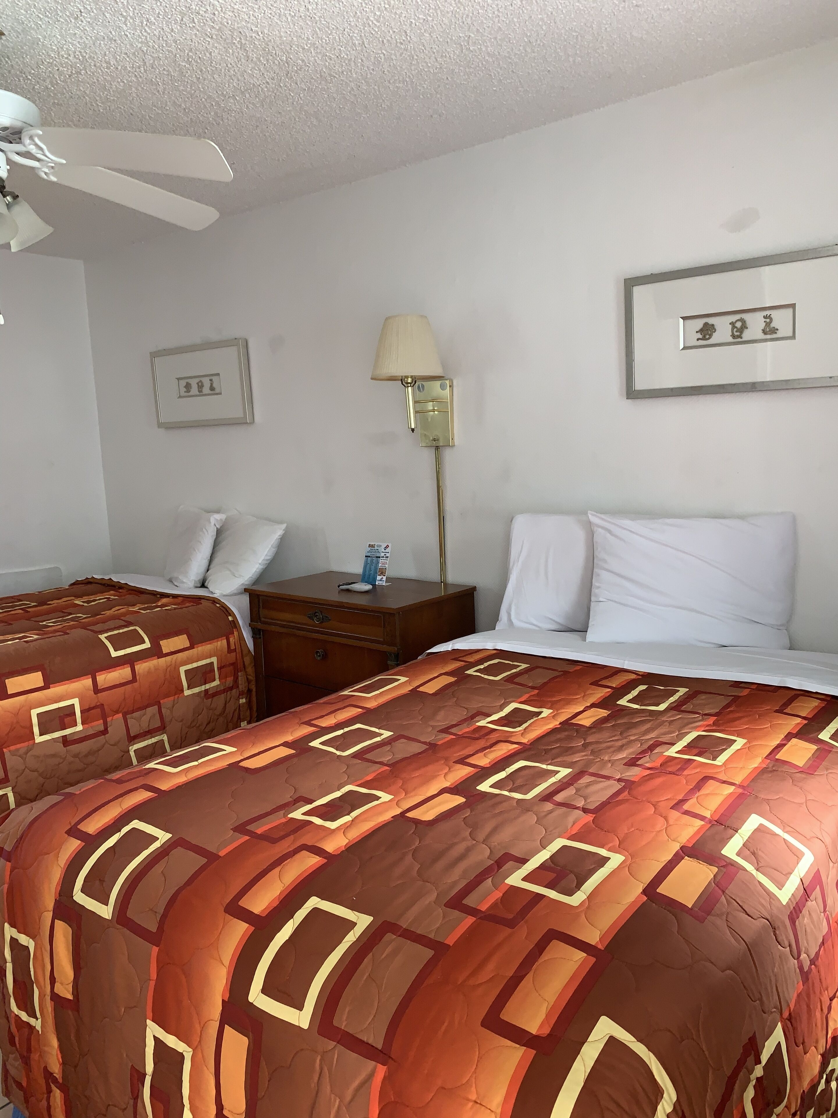 Una habitación de hotel con dos camas, un ventilador de techo, una mesita de noche con lámpara y cuadros en la pared.