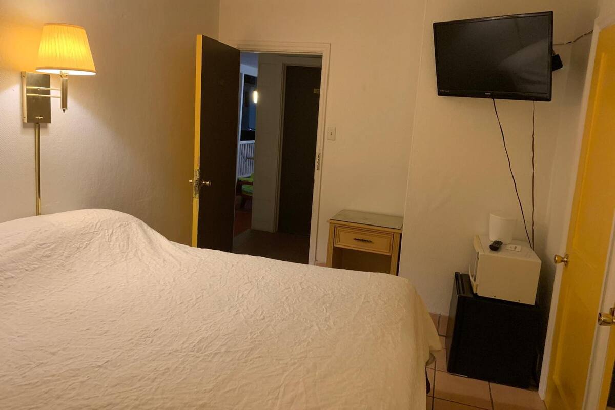 A hotel room with a bed, a bedside lamp, a television mounted on the wall, and a door leading to another room.
