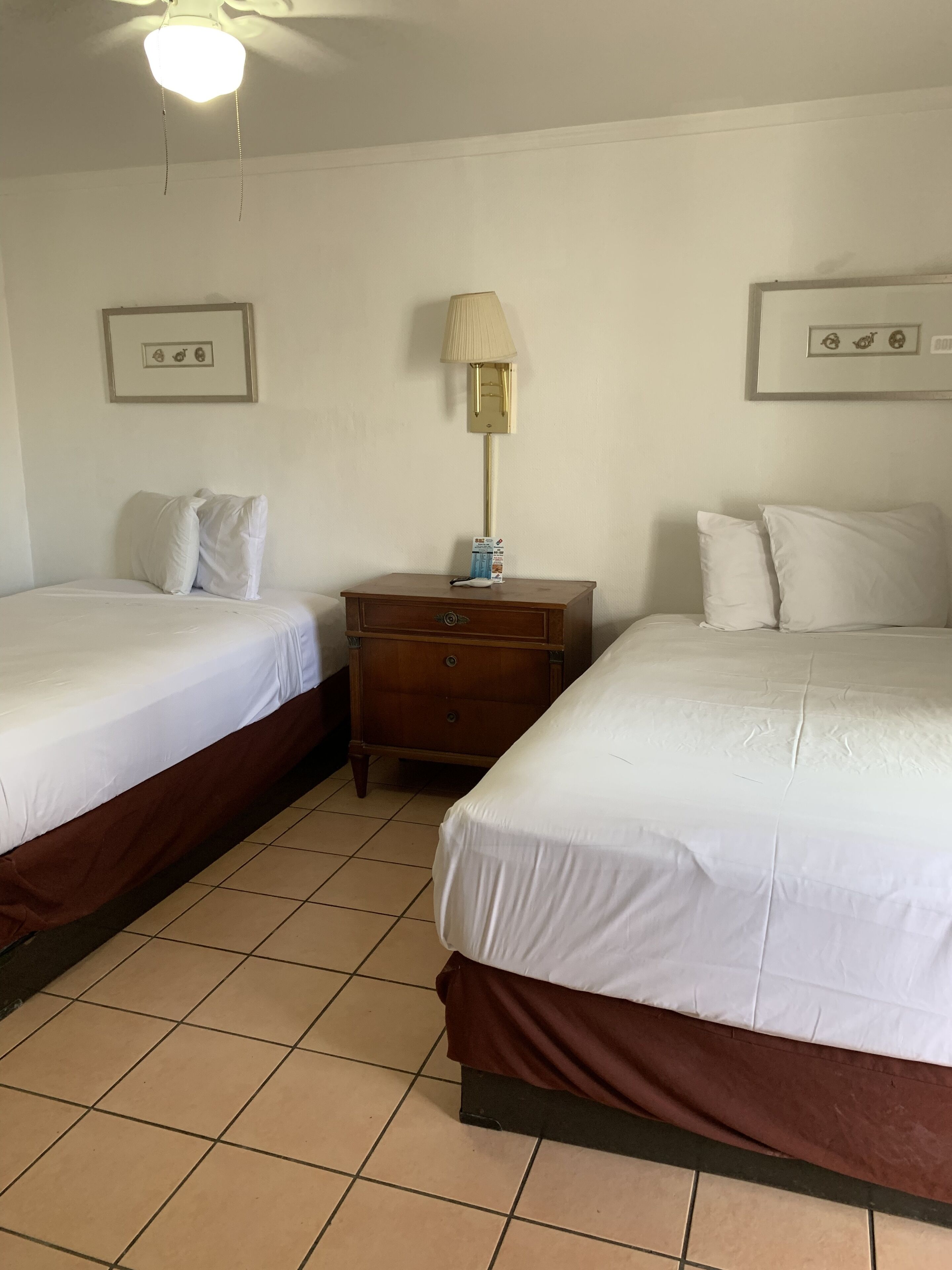 A hotel room with two single beds, a wooden nightstand, a lamp, and framed pictures on the walls.