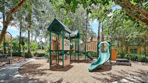 Ein Spielplatz mit Rutsche und Klettergerüsten, umgeben von Bäumen und einer gepflasterten Fläche.