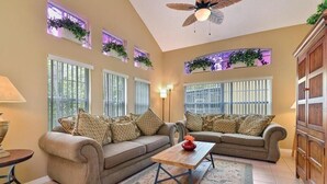 Ein Wohnzimmer mit zwei Sofas, einem Couchtisch und einer Lampe. Es gibt Fenster mit Pflanzen und einen Deckenventilator.