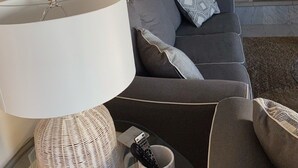 Un salon avec un canapé gris, une lampe blanche, une table d’appoint en osier et un téléviseur fixé au mur.