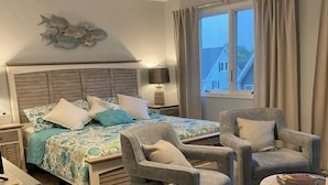 Une chambre à coucher comprenant un lit, deux fauteuils, un pouf rond, un ventilateur de plafond, une table de chevet et une fenêtre avec des rideaux.