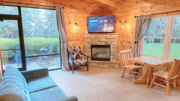 Un acogedor interior de cabaña con chimenea de piedra, un televisor montado en la pared, un sofá y una mesa de comedor con sillas.