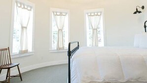 A bedroom with a curved ceiling, a bed with a black frame, a wooden chair, and three windows with curtains. - Unique Silo Suite with Jacuzzi Tub, Kitchen, Fireplace, Living Room (Millersburg)