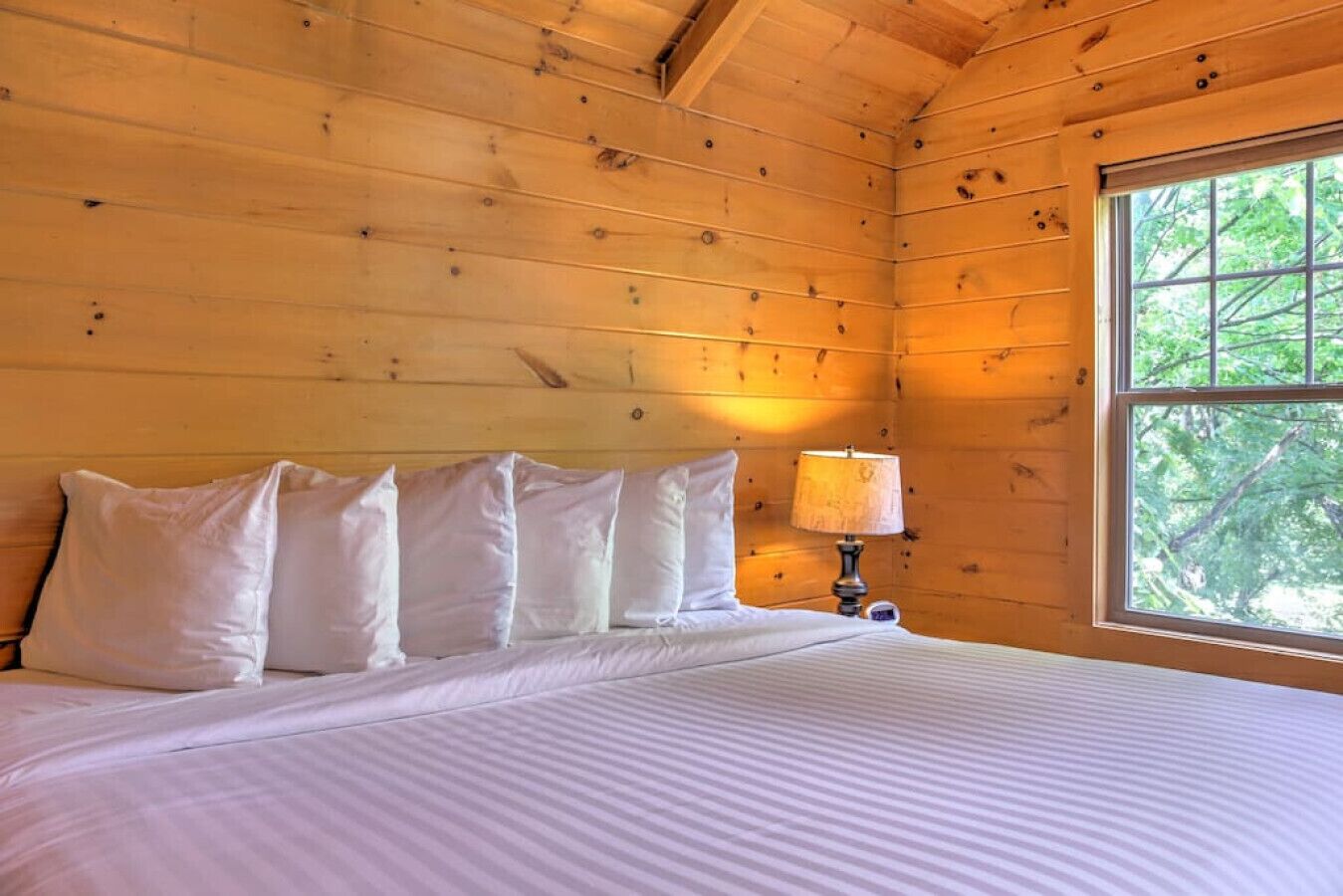 A wooden cabin room with a bed, pillows, a nightstand, and a window with a view of trees.