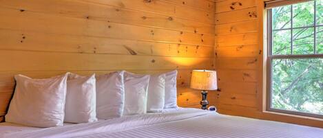 A wooden cabin room with a bed, pillows, a nightstand, and a window with a view of trees.