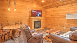 Interior acogedor de una cabaña de madera con chimenea, sillones de cuero, una mesa redonda de madera y un televisor montado.