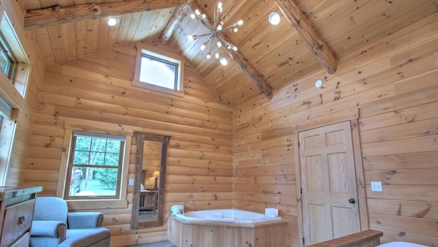 Un dormitorio tipo cabaña de troncos con techo de madera, una ventana grande, una bañera integrada, una cama y una silla.
