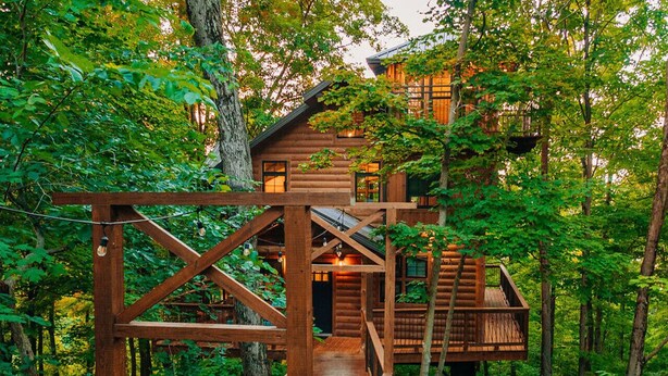 Una cabaña de madera con un balcón de madera y escaleras que conducen al edificio principal, rodeada de un bosque denso.