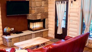 Een gezellig chaletinterieur met een stenen open haard, een flatscreen televisie aan de muur, een rode leren bank en een houten bijzettafeltje.