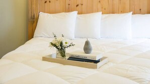 Un lit bien fait, avec du linge de lit blanc, un vase rempli de fleurs blanches et un livre intitulé « Sel pour la mer » posé sur un plateau.