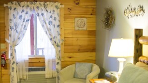 A cozy room with a wooden interior, a bed, a chair with a cushion, a bedside table with a lamp, and a window with curtains. - Romantic Couples Suite with Jacuzzi Tub and Kitchenette (Berlin)