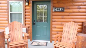 Una cabaña de madera con puerta verde, dos sillas Adirondack de madera y un felpudo.