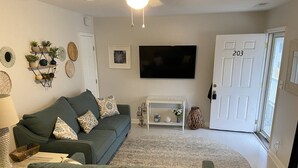 En stue med en grå sofa, et fladskærms-tv, en hvid dør med nummer 203, og et lille hvidt konsolbord med pynt.