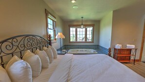 A large bed with white linens, a window with blinds, a small table with towels, and a bathtub.