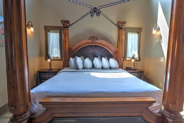 A large bed with a wooden headboard, flanked by two nightstands with lamps, in a room with wooden columns and a large window with curtains.