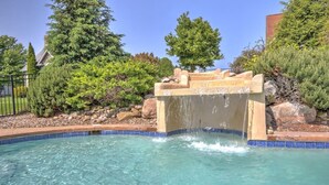 Piscina translúcida com cachoeira de pedra, cercada por um jardim paisagístico com arbustos e árvores diversos.