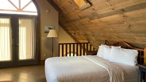 Quarto com teto de madeira, cama com roupa branca, mesa de cabeceira, abajur e vista para o exterior através de uma porta com cortinas.