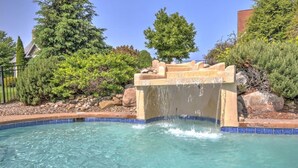 Piscina límpida com uma cascata de pedra, rodeada por vegetação ajardinada e uma grade de metal.