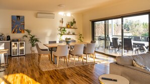 A modern living room with a wooden floor, a dining area with a wooden table and chairs, a sofa, and a large sliding glass door leading to an outdoor patio.