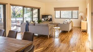 A spacious living area with a sofa, a coffee table, and a TV. There is a dining table with chairs and a balcony with a railing.