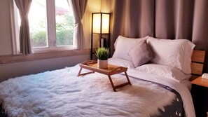 A bedroom with a large bed, a bedside table with a lamp and a plant, and a window with curtains.