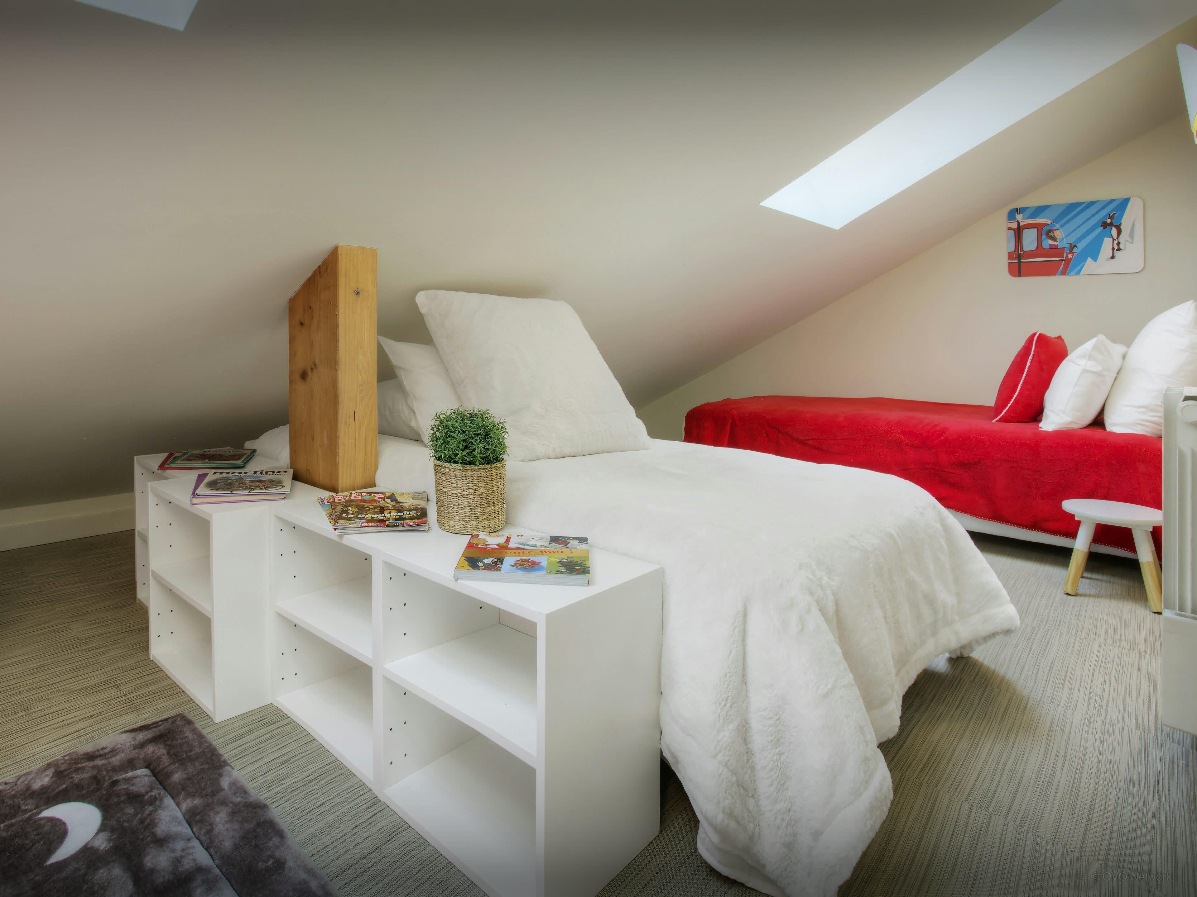 A bedroom with a sloped ceiling, a bed with a red cover, white pillows, a small white side table with books and a plant, and a skylight.