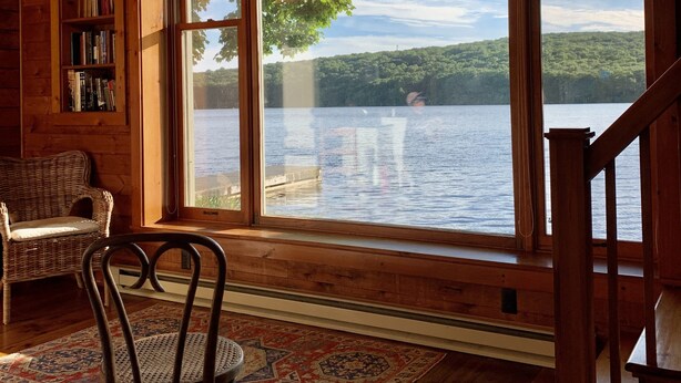 A wooden cabin with a large window overlooking a lake, a wicker chair, a bookshelf, and a patterned rug. - Rustic Lakeside Cabin at White Gate Farm (East Lyme)