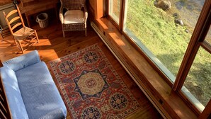 A cozy cabin interior with a large window, wooden walls, a blue checkered sofa, a wicker chair, and a patterned rug. - Rustic Lakeside Cabin at White Gate Farm (East Lyme)