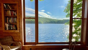 A room with a large window offering a view of a lake and forest. There is a bookshelf and two wicker chairs. - Rustic Lakeside Cabin at White Gate Farm (East Lyme)