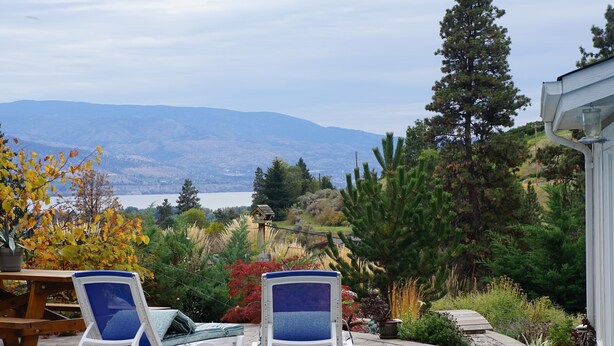A patio with two lounge chairs, a wooden table, and a view of mountains and a lake. - Dreams B&B (Summerland)