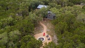 Vista aerea di una struttura circondata da fitta vegetazione, con un piccolo edificio a tetto spiovente e un vialetto lastricato che conduce a una zona salotto circolare con sedie.