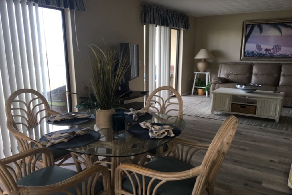 A dining area with a glass table surrounded by wicker chairs, a television, and a sofa in the background.