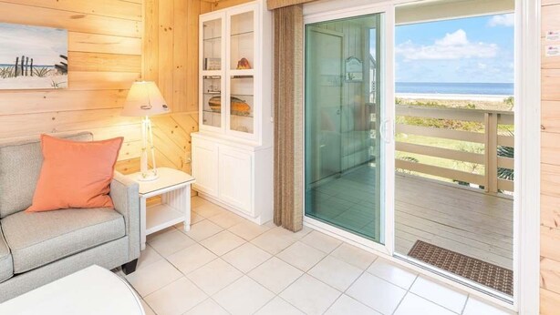 A room with a sofa, a small table with a lamp, a glass door leading to a balcony with a view of the beach. - Lighthouse Point 30C | Ocean View Condo, Pools! (Tybee Island)