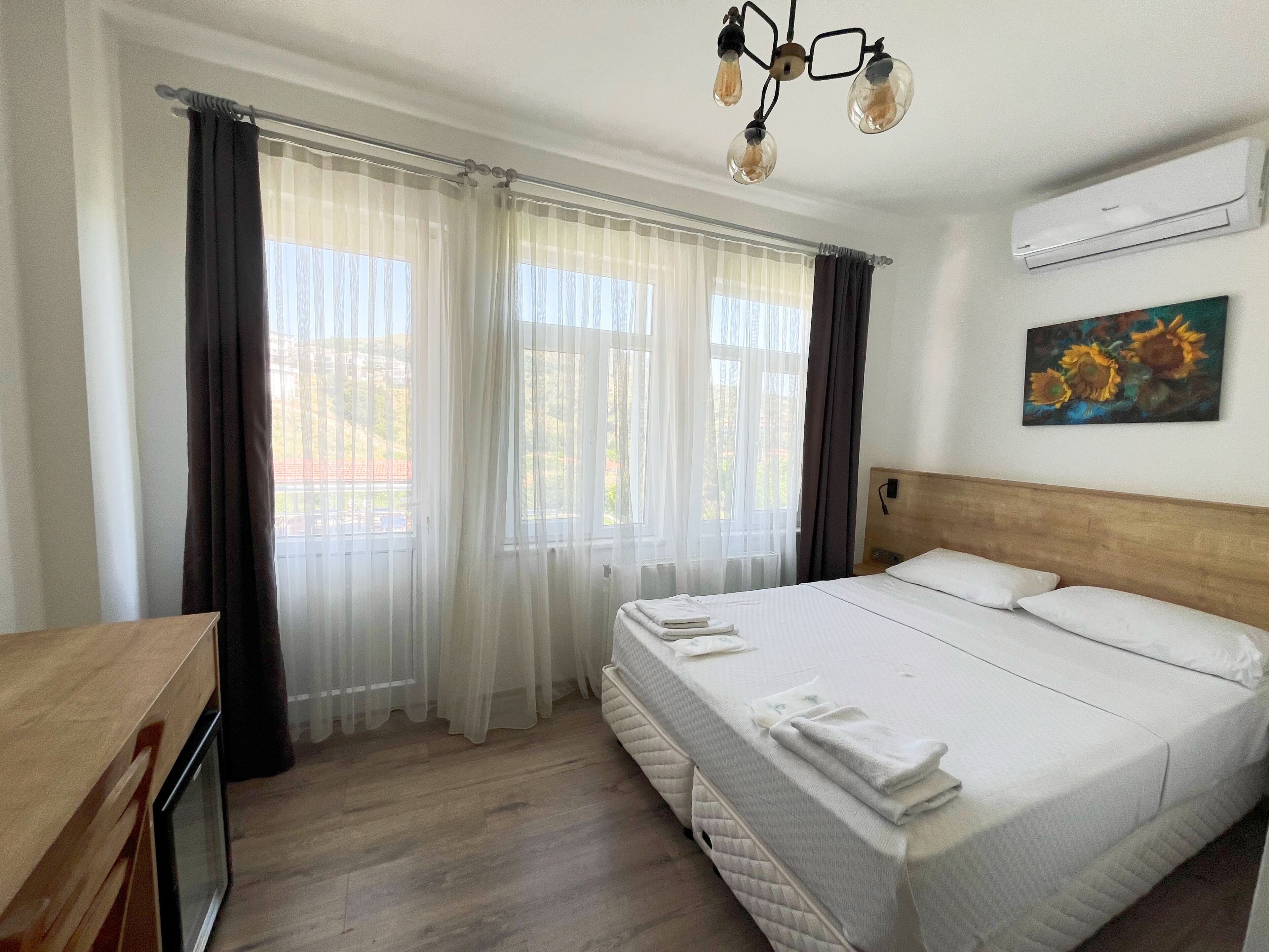A hotel room with a bed, curtains, a painting, an air conditioner, and a wooden desk.