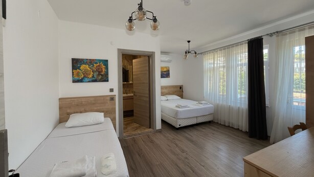A hotel room with a wooden floor, a bed with white linens, a desk, a chair, a painting on the wall, and a door leading to a bathroom. - Parli Hotel (Gökçeada)
