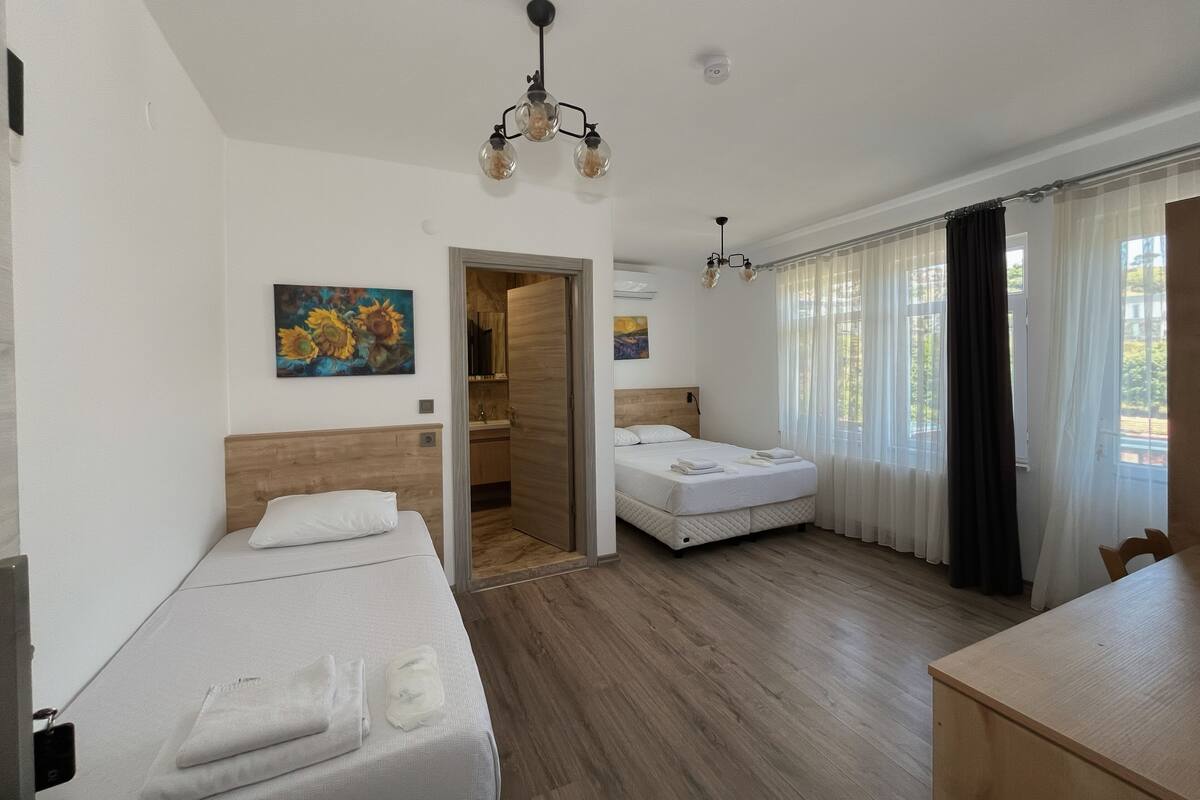 A hotel room with a wooden floor, a bed with white linens, a desk, a chair, a painting on the wall, and a door leading to a bathroom.
