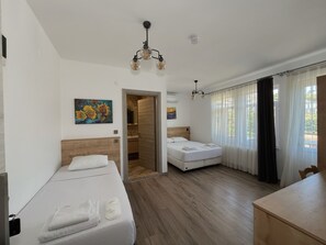 A hotel room with a wooden floor, a bed with white linens, a desk, a chair, a painting on the wall, and a door leading to a bathroom. - Parli Hotel (Gökçeada)