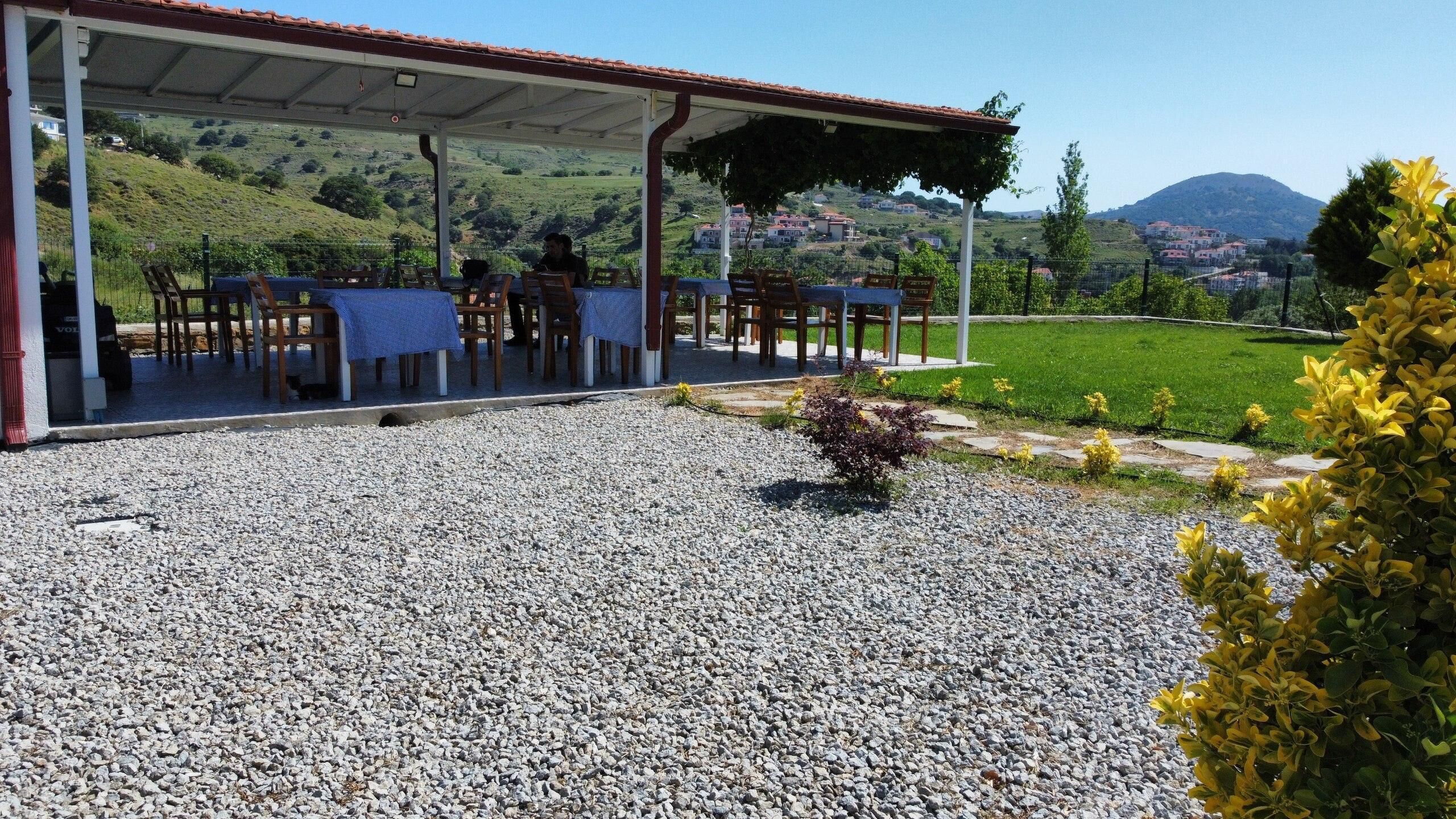 Un espace extérieur abrité, aménagé avec des tables et des chaises, doté d’un sol en gravier et offrant une vue sur un paysage verdoyant avec des collines et des bâtiments à l’horizon.