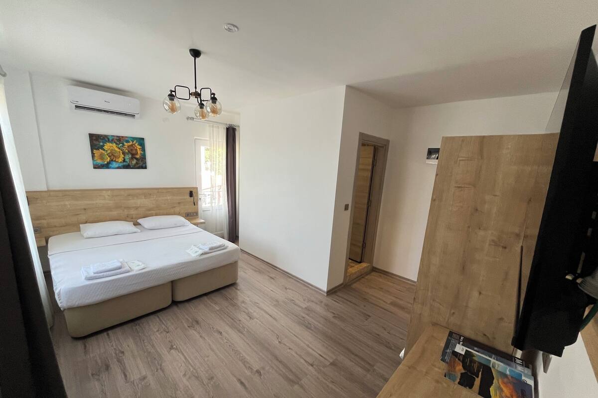A hotel room with a wooden floor, a bed with white bedding, a painting on the wall, and a ceiling light fixture.