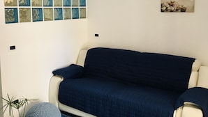A room with a blue quilted sofa, a white wall with a grid of blue and green glass panels, and two framed pictures depicting a beach scene and a cityscape. - APARTMENT IN VILLA (SANGINETO)